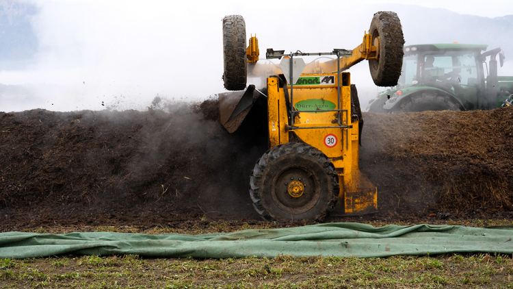 Eine dampfende Mistmiete wird mit Hilfe von einem Kompostwender mechanisch gewendet.