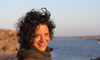 Cellistin Barbara Gasser blickt in die Kamera. Im Hintergrund ist verschwomen Wasser zu erkennen. 