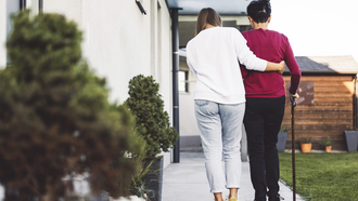 Eine junge Frau stützt eine Person, welche an einem Gehstock geht