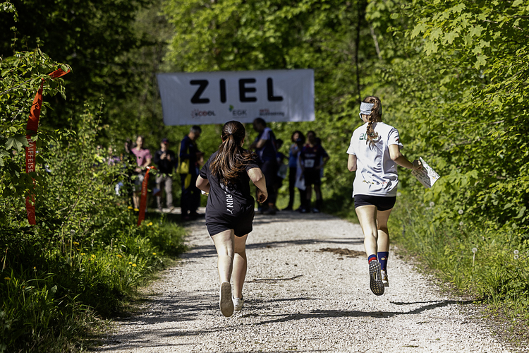 Zwei Läuferinnen rennen auf einem Waldweg auf ein „Ziel“-Banner zu, im Hintergrund warten Zuschauer am Rand der Strecke. Zwei Läuferinnen rennen auf einem Waldweg auf ein „Ziel“-Banner zu, im Hintergrund warten Zuschauer am Rand der Strecke.