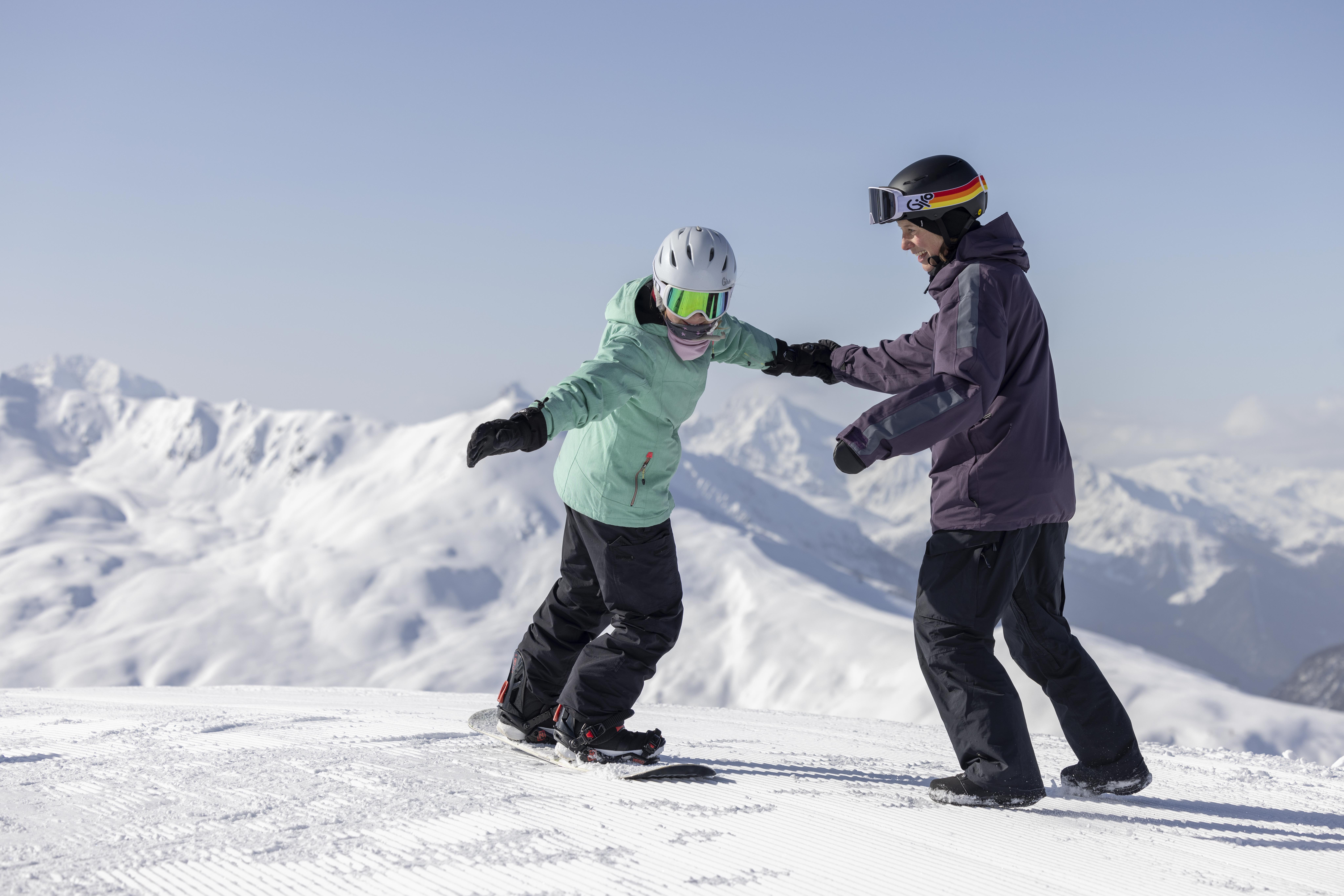 Zwei Personen im Schneesportlager Davos beim Snowboarden auf einer verschneiten Piste, umgeben von den verschneiten Alpen unter blauem Himmel.