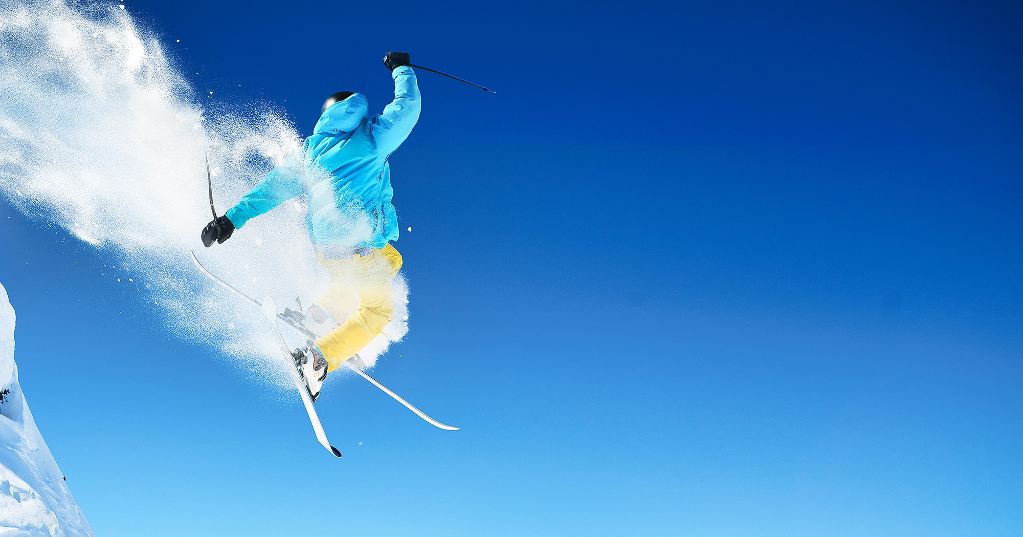 Skifahrer in blauer Jacke und gelber Hose springt mit Skiern durch die Luft, während Schneepulver aufwirbelt, vor klarem, blauem Himmel.