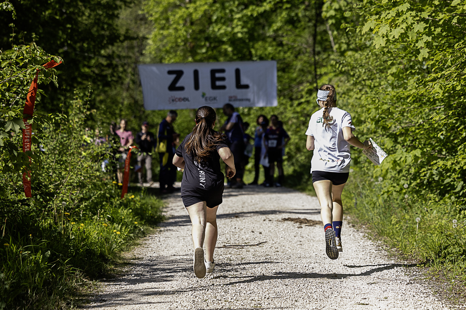 Zwei Läuferinnen rennen auf einem Waldweg auf ein „Ziel“-Banner zu, im Hintergrund warten Zuschauer am Rand der Strecke.